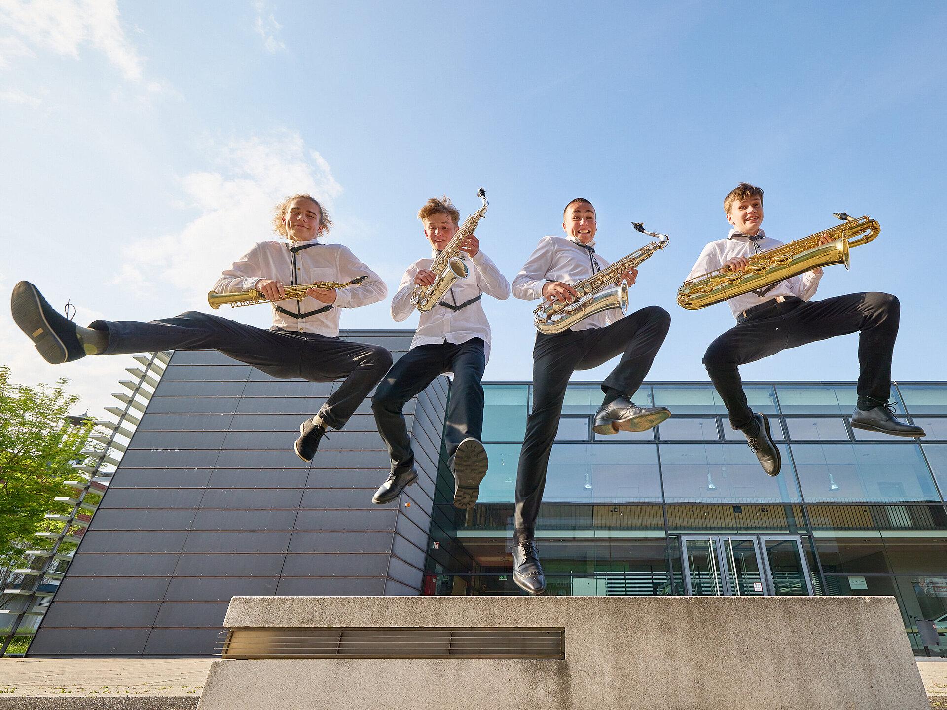 Vier Jugendliche mit je einem Saxophon in der Hand springen in die Luft.